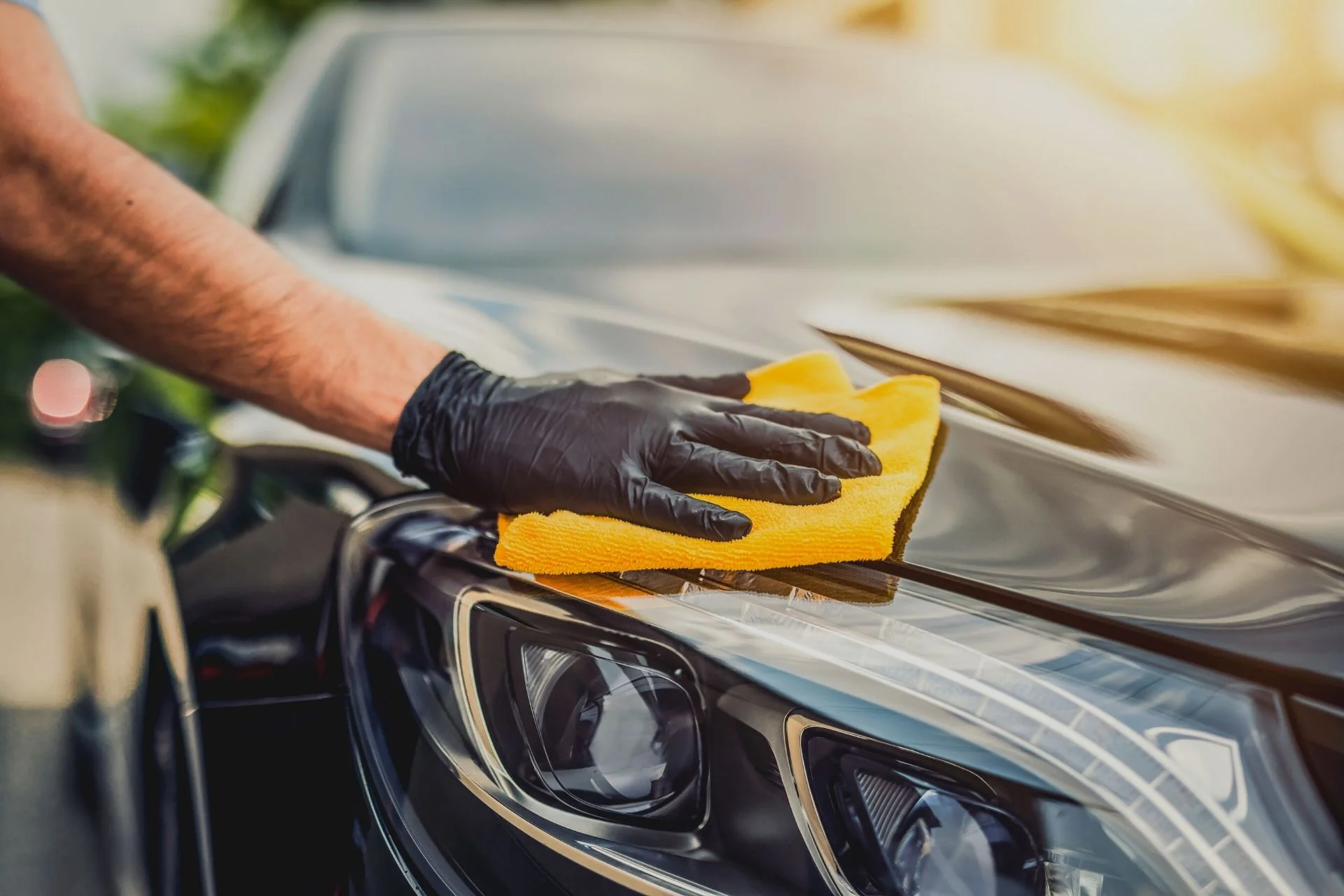 A professional detailer wearing black gloves using a yellow microfiber cloth to wipe and polish the hood and headlight of a luxury car, featuring exterior ceramic coating and window tinting maintenance.