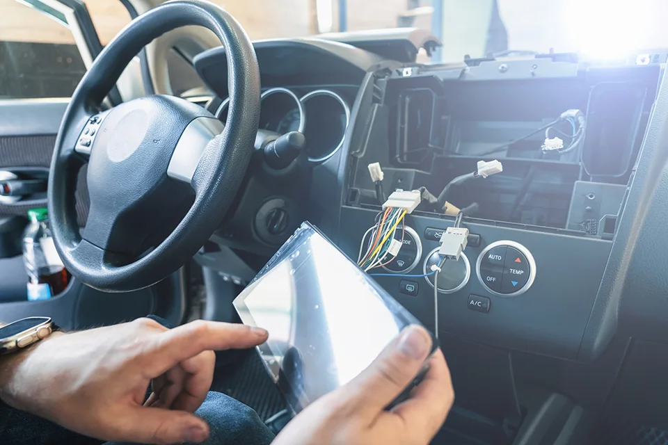 A mechanic holding a digital touchscreen display unit while working on the dashboard and wiring of a car stereo installation.